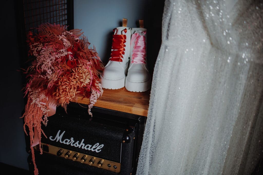 White doc martens sitting on top of a large Marshall amp, between a hanging sequin-covered wedding dress and red and pink dried flower bouqet
