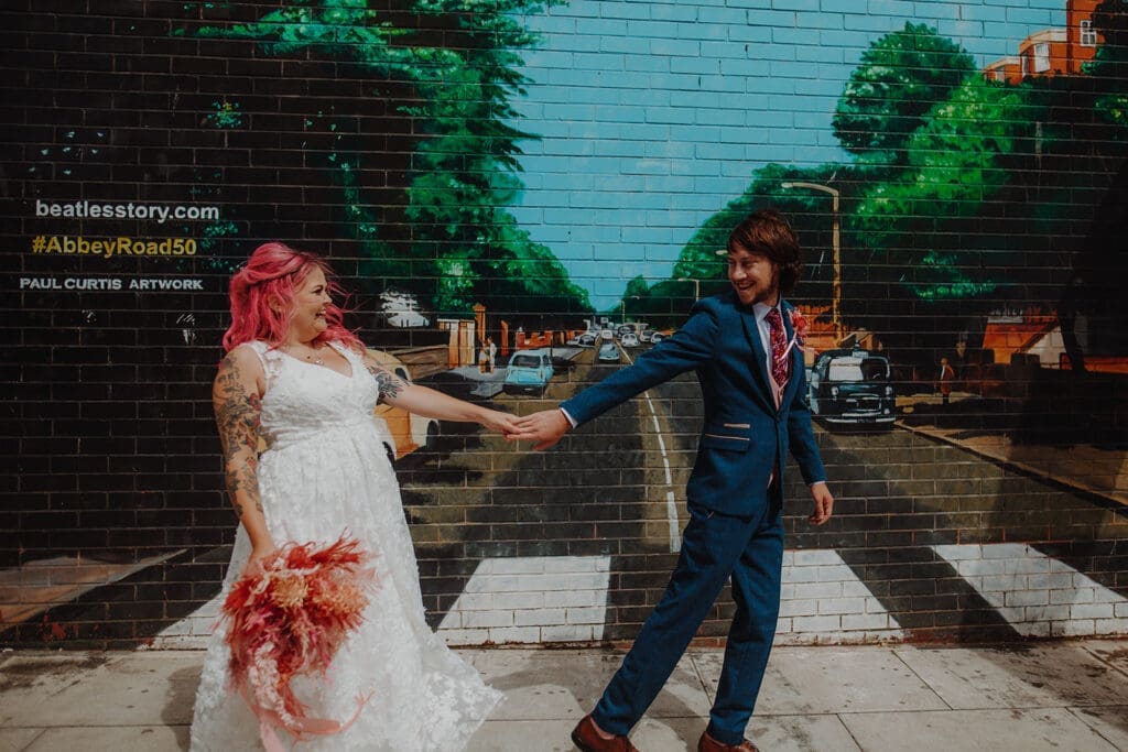 The groom leads his bride over a mural of Abbey Road. Both are laughing and the bride holds a bouquet in muted pinks and reds.