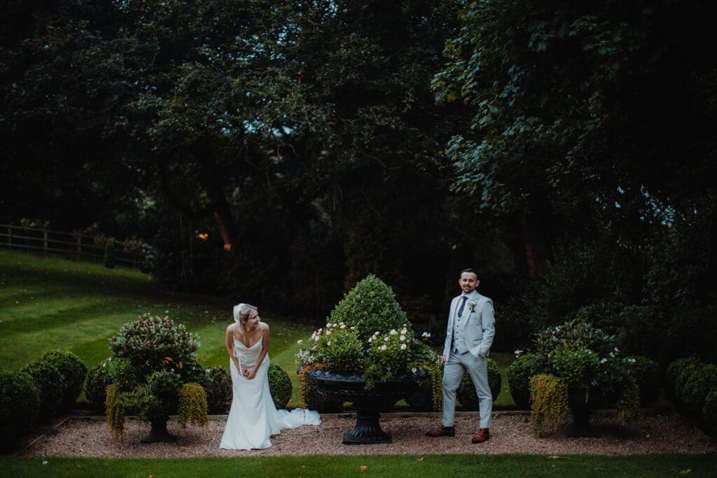 The bride leans forward to see her new spouse on the other side of the statue and foliage between them.