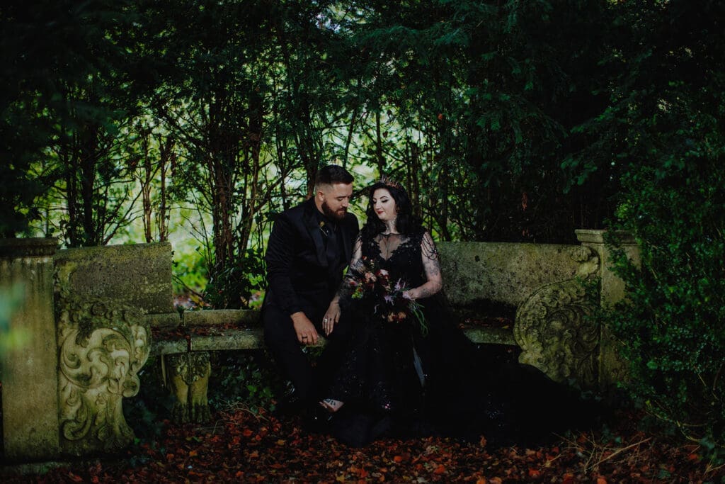 The bride and groom sit on a mossy stone bench in the middle of a forest, surrounded by greenery and on a floor of autumn leaves. The bride is wearing a black wedding dress and gold spiked crown. The groom dons an all-black suit and tie.
