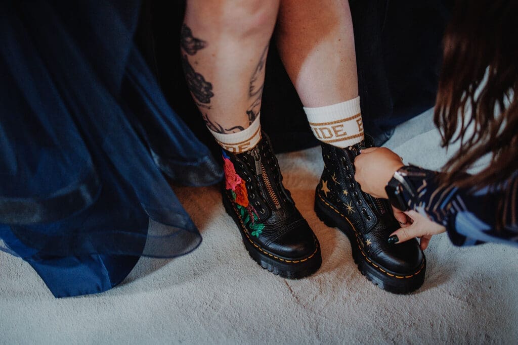 A bride getting her Doc Martens zipped up. Peeping over the top of her docs are socks that read "BRIDE". Her docs have embroidered roses and stars on. 