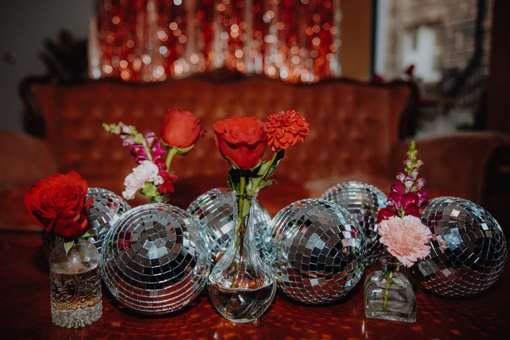 Six disco balls placed in a line on a table, with little vases holding red roses and pink peonies. The background shows red vintage furniture and glittering sequin backdrops.