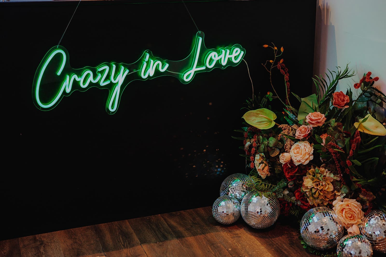 A neon sign that reads 'crazy in love'. Below it are a number of different sized disco balls and an arrangement of flowers featuring different shades of green, red and orange.