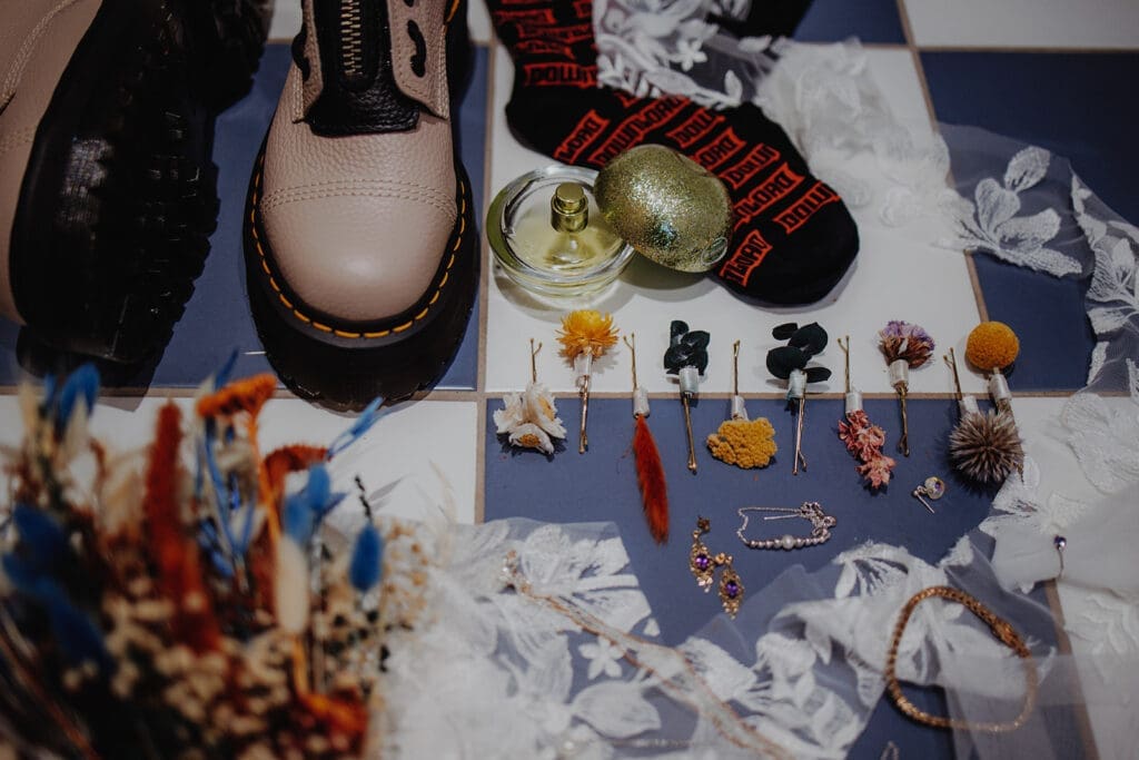 A flay-lay of lots of wedding items. It includes colourful button holes of yellow, red and blue, jewellery laid out, a pair of Doc Martens, Download Festival socks, perfume, and a bouquet of dried flowers that include blue, orange and yellow, as well as beautiful white lace draped over the table.