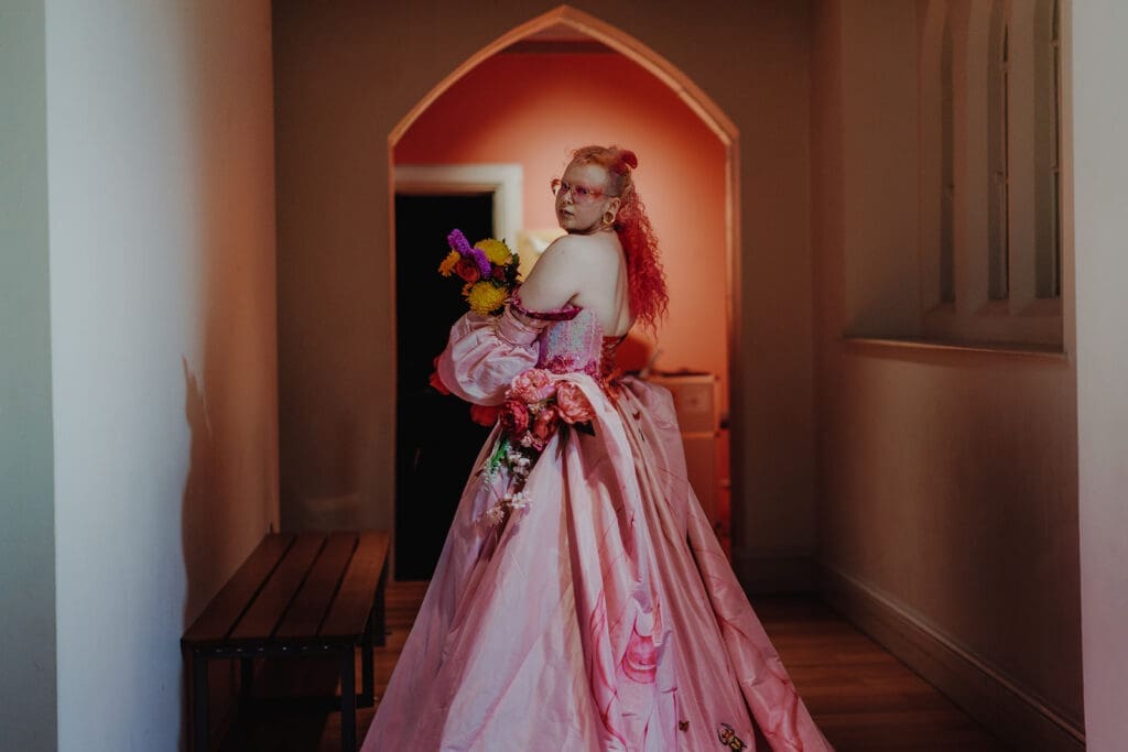 A gorgeous nearly-wed stands in an arched entry way, showing off the back of their pink corseted dress. Their long pink hair is half up half down, and they're looking over their shoulder to the camera.