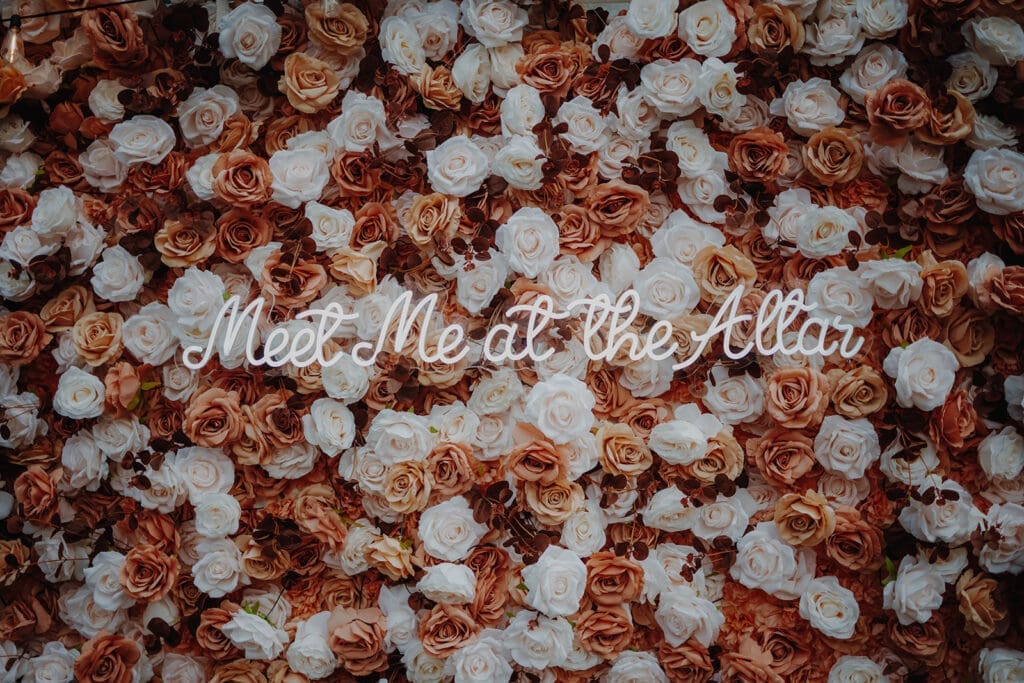 A flower wall made up of white, pink and mocha coloured roses and a neon light in the middle that reads "Meet me at the altar"