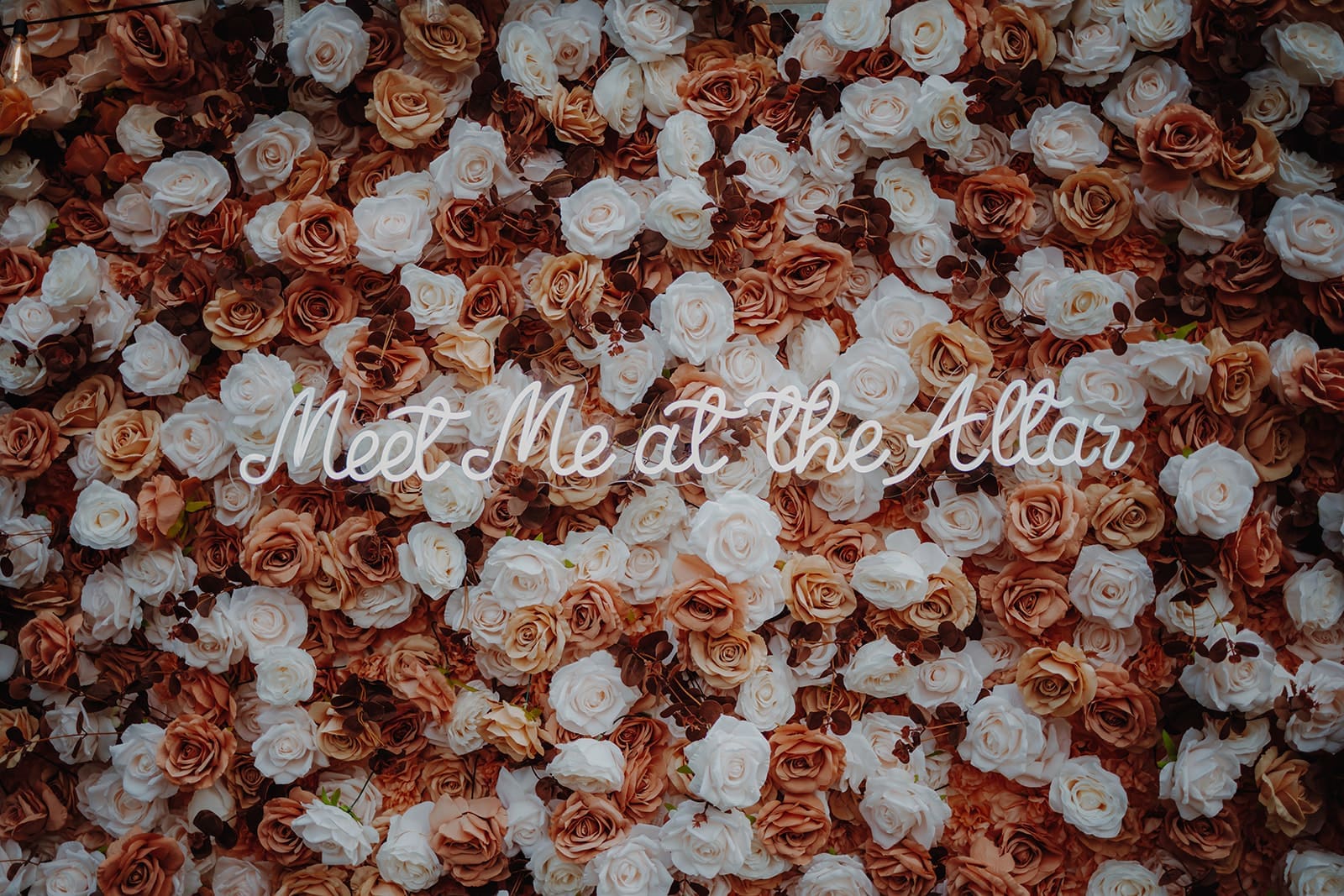 A flower wall made up of white, pink and mocha coloured roses and a neon light in the middle that reads "Meet me at the altar"