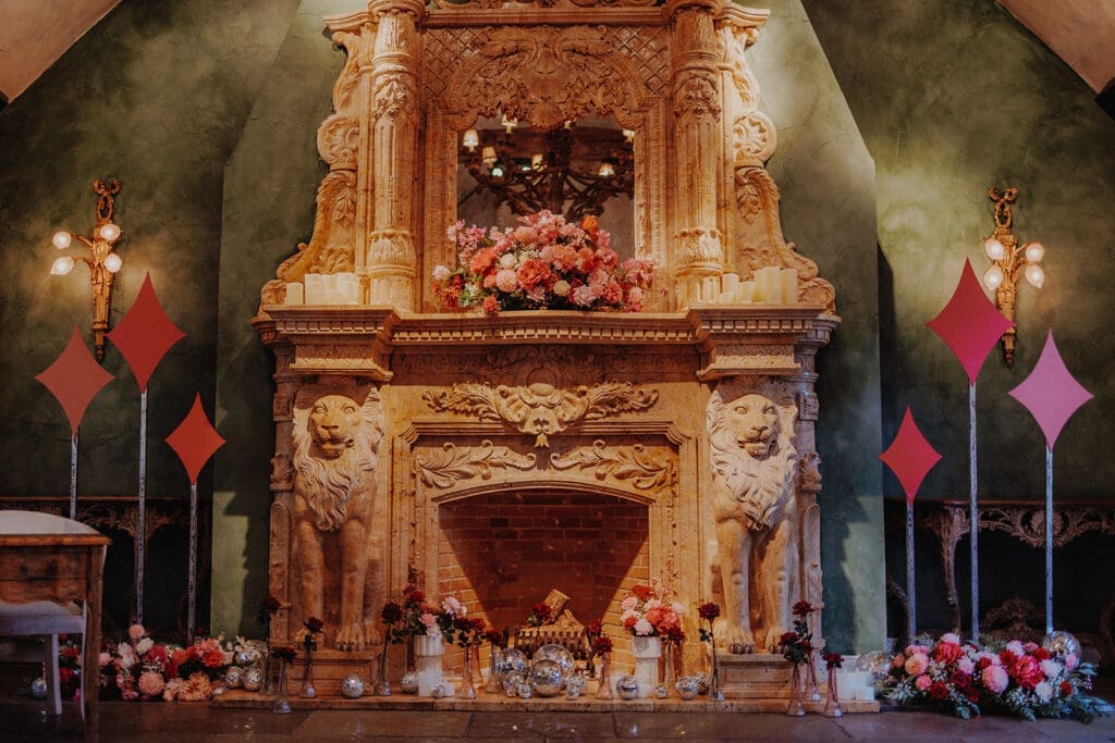 A large, grand fireplace with a floral display of pink and red roses. Below the fireplace are scattered disco balls and vases of peonies and roses.